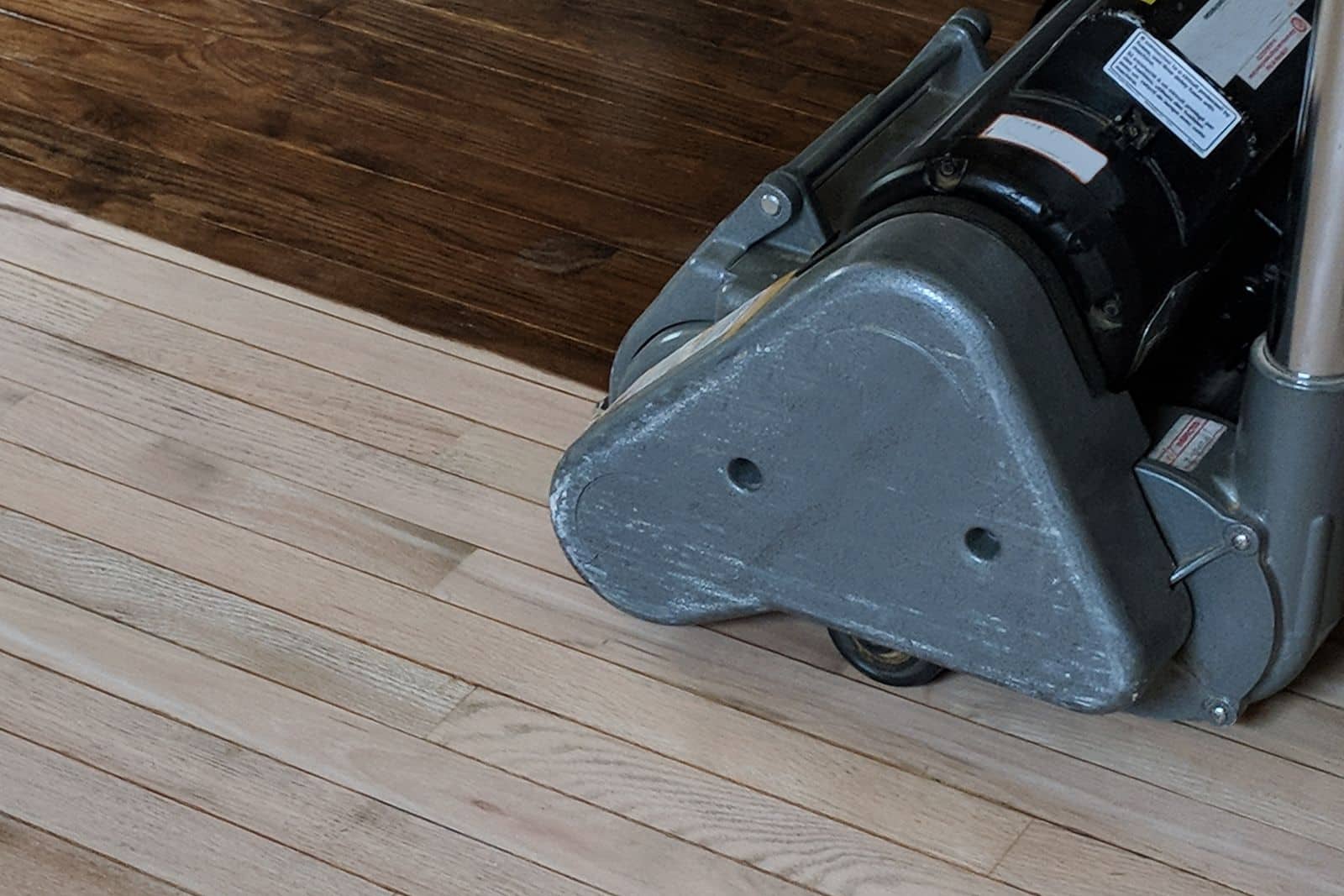 Drum sander on an oak strip floor, half sanded to bare wood and half still in the old dark finish