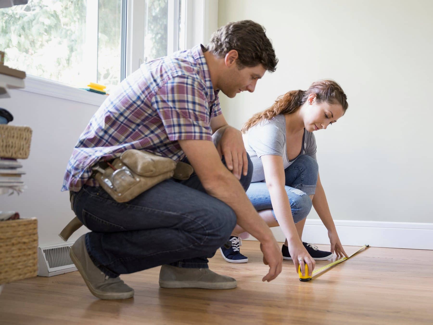 Two people kneeling on a hardwood floor, measuring the room with a tape measure